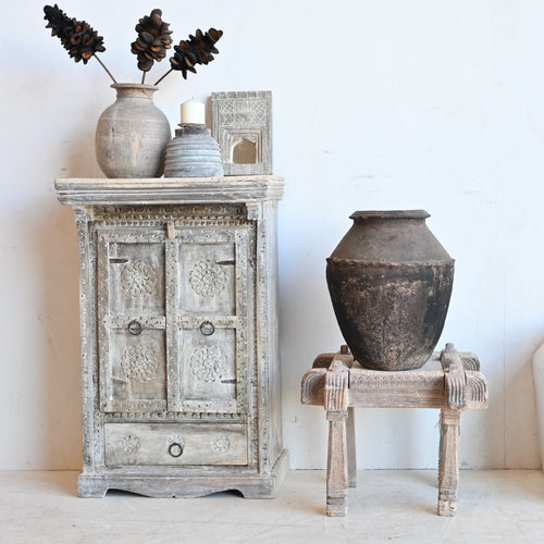 A rustic carved wooden cabinet with a distressed gray finish stands against a white wall, styled with a ceramic vase of pinecones, a candle, and a small box. In the center, a large dark earthenware pot sits on a small wooden stool, and to the right is a white upholstered bench with a cushion.