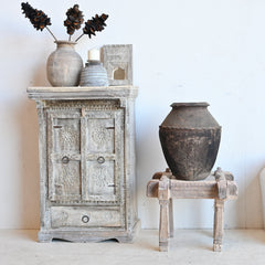 A rustic carved wooden cabinet with a distressed gray finish stands against a white wall, styled with a ceramic vase of pinecones, a candle, and a small box. In the center, a large dark earthenware pot sits on a small wooden stool, and to the right is a white upholstered bench with a cushion.