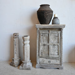 A distressed, light gray carved wooden cabinet styled with a dark brown ceramic pot and a small gray vase on top, next to two matching carved pillar candle holders against a plain off-white wall.