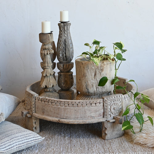 A rustic carved wooden decorative tray holding two candlesticks and a small potted green plant, styled on a woven rug with pillows against a white wall.