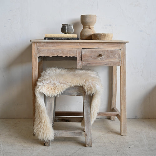 A rustic light wood writing desk with a single drawer, styled with a fluffy cream sheepskin stool cover, books, and small wooden decor objects against a textured off-white wall.