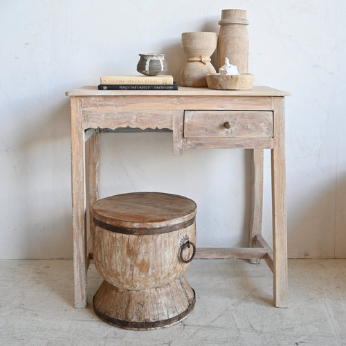 A rustic light wood writing desk with a single drawer and a small wooden stool in front, styled with ceramic vases and books against a white wall.