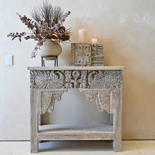 A distressed light gray carved wooden console table styled with a ceramic vase of dried botanicals and candles against a neutral beige wall.