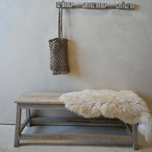 A rustic wooden bench with a fluffy cream sheepskin throw sits against a textured beige plaster wall. Above the bench, a weathered gray wooden peg rail is mounted on the wall, holding a woven brown tote bag.