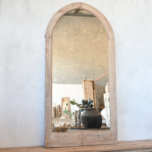 An arched wooden wall mirror with a natural light wood frame, mounted on a textured white plaster wall above a carved wooden mantel. The mirror reflects a home interior 