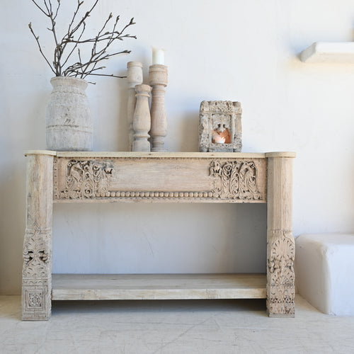 Decorative wooden console table with carved details, candles, and a vase against a white wall.